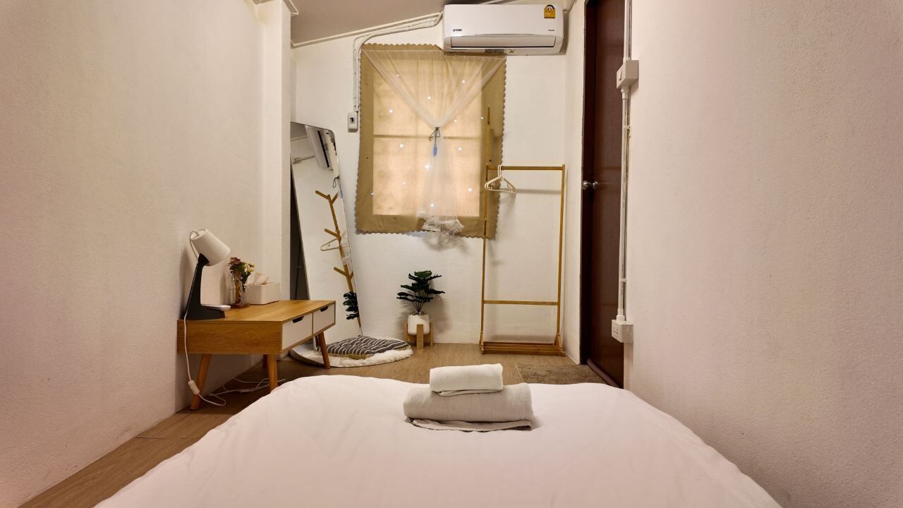 A neatly arranged hotel room with a bed, a desk, a small plant, a window with curtains, and an air conditioning unit.
