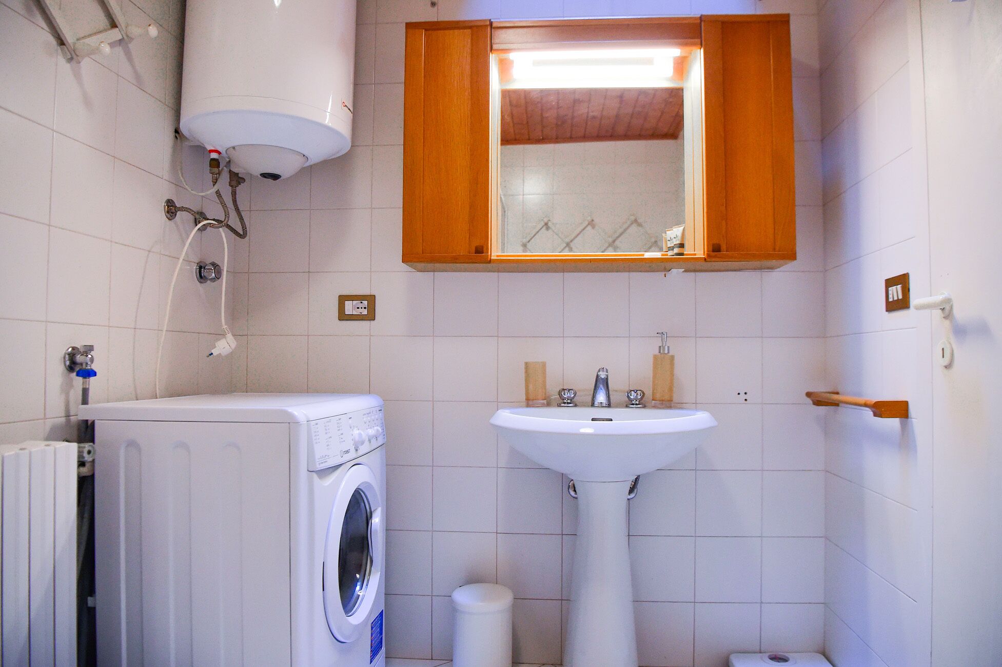 A compact bathroom with a washing machine, a sink, and a wooden cabinet above the mirror.
