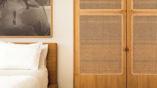 A bedroom with a bed, white pillows, and a wooden headboard. There is a wooden wardrobe with a woven door and a framed picture on the wall above the bed.