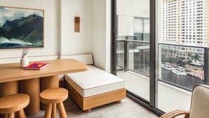 Une pièce moderne avec une table à manger en bois, un banc rembourré et deux tabourets. Une grande fenêtre offre une vue sur le paysage urbain et l’étendue d’eau.