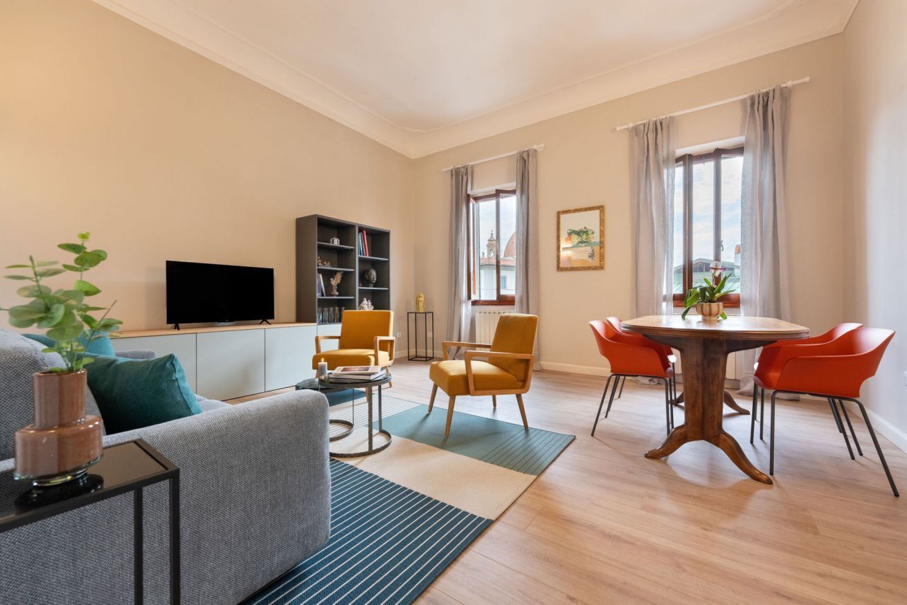 A living room with a sofa, a TV, a bookshelf, a dining table with chairs, and a window with curtains.
