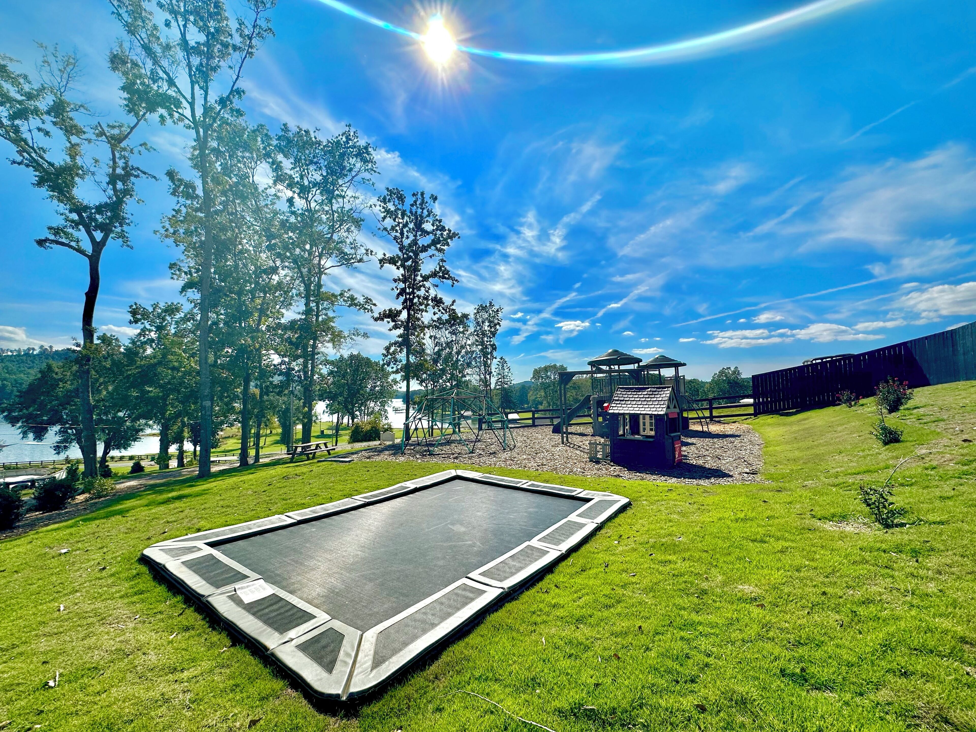 A trampoline on a grassy field with trees and a small playhouse in the background.