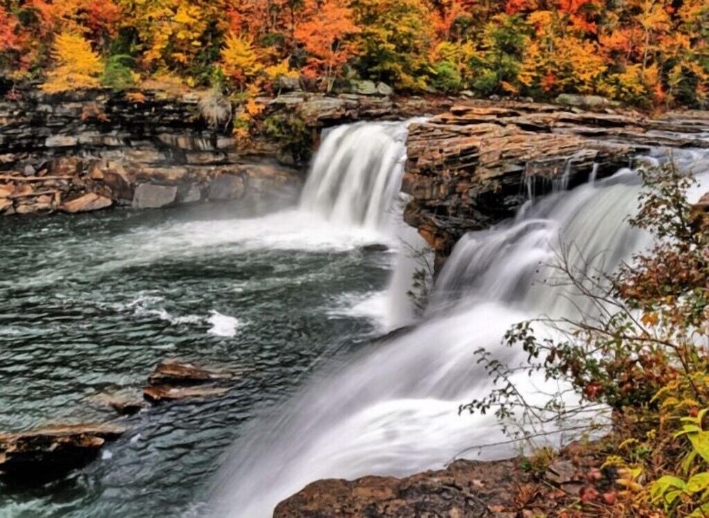 Une cascade d’eau se déversant sur un terrain rocheux, avec le feuillage automnal en arrière-plan.