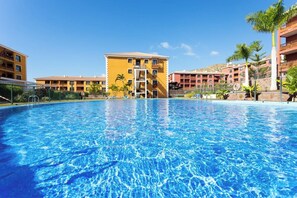 A swimming pool surrounded by yellow buildings with balconies, a clear blue sky, and palm trees.