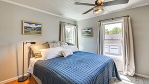 Quarto com cama, ventoinha no teto e duas janelas com cortinas.