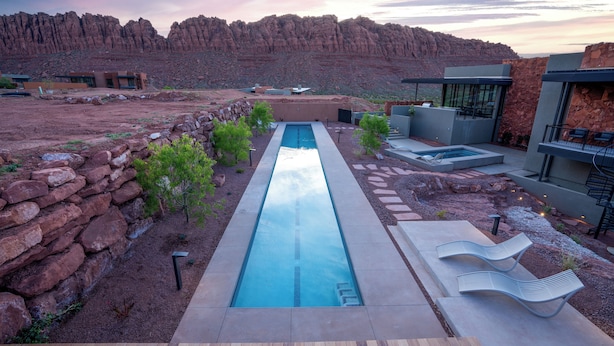 Un área de piscina moderna al aire libre con una piscina rectangular larga, sillas de descanso y un muro de piedra.