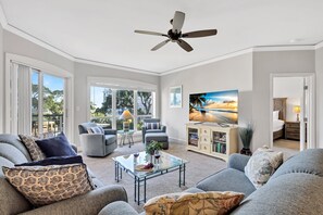 A spacious living room with a ceiling fan, a glass coffee table, grey sofas, and a large window offering a view of the outdoors.