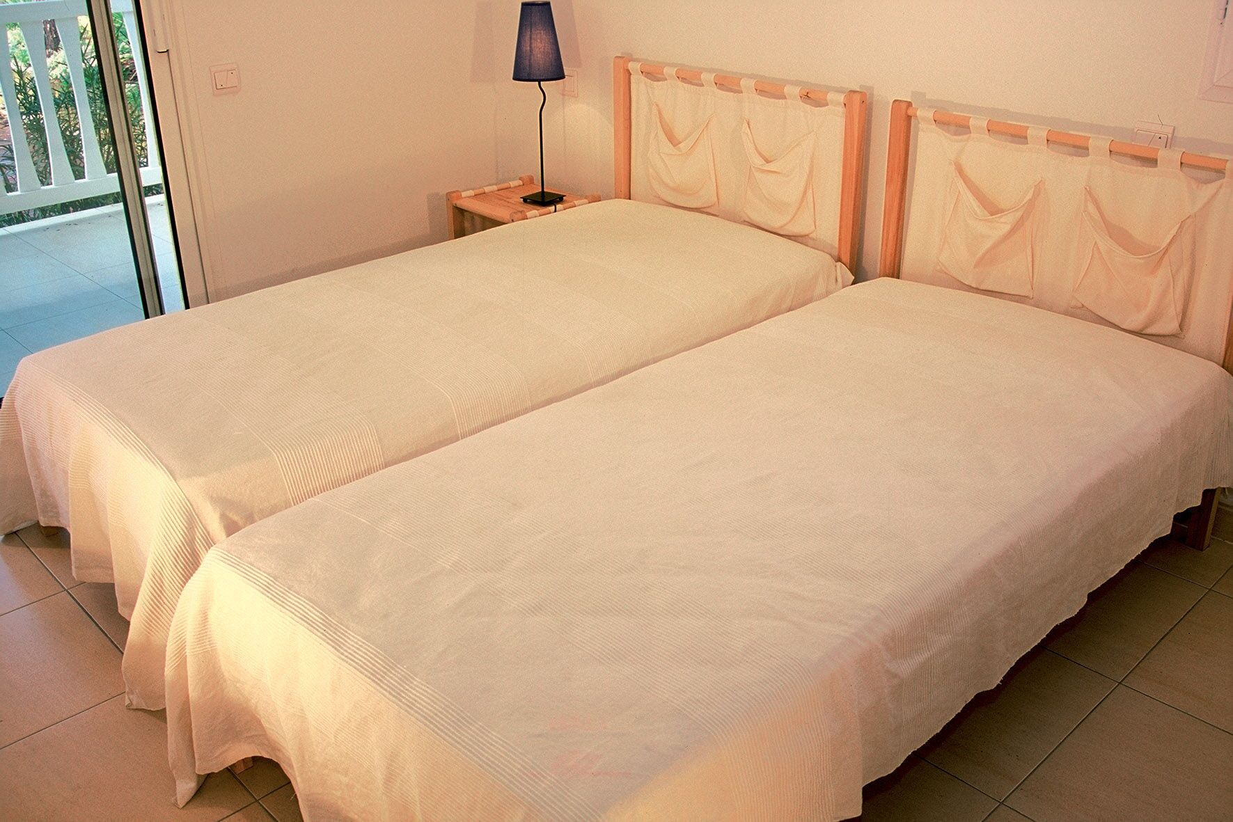 A room with two single beds, a nightstand with a lamp, and a view of the outside balcony.