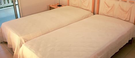 A room with two single beds, a nightstand with a lamp, and a view of the outside balcony.
