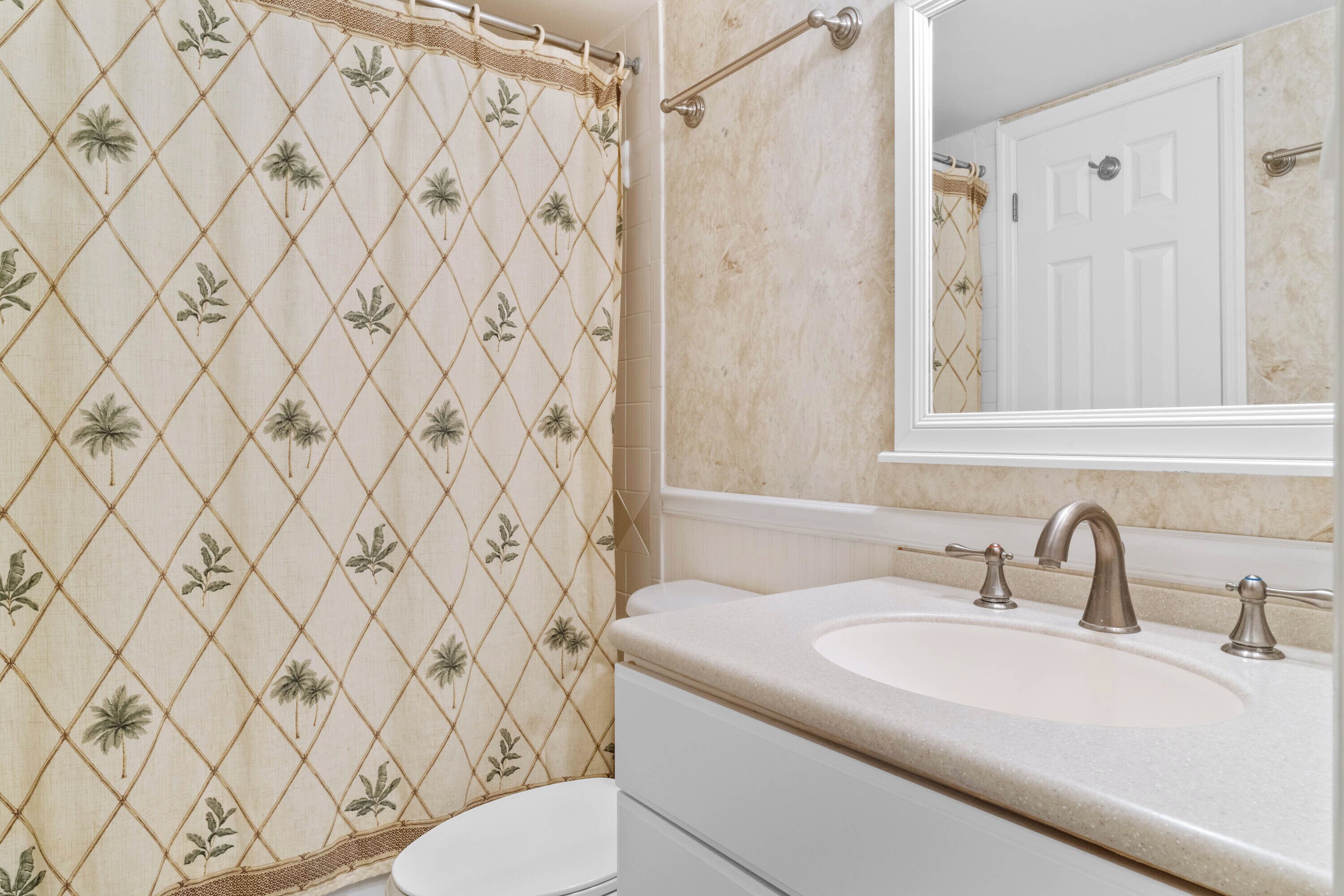 Un baño con cortina de ducha estampada con diseño de rombos, un inodoro blanco, un lavabo con un solo grifo y un espejo que refleja una puerta blanca.