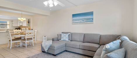 Una sala de estar con un sofá seccional gris, un comedor con mesa y sillas blancas, un ventilador de techo y un cuadro con una escena de playa.