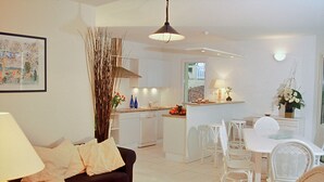 Una cocina moderna con armarios blancos, un comedor con sillas blancas y una sala de estar con un sofá oscuro y una lámpara.