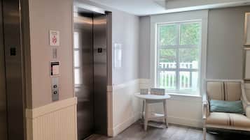 Een lift met een bord waarop noodprocedures staan, een kleine tafel met motiverende bordjes, en een raam met uitzicht op groen.