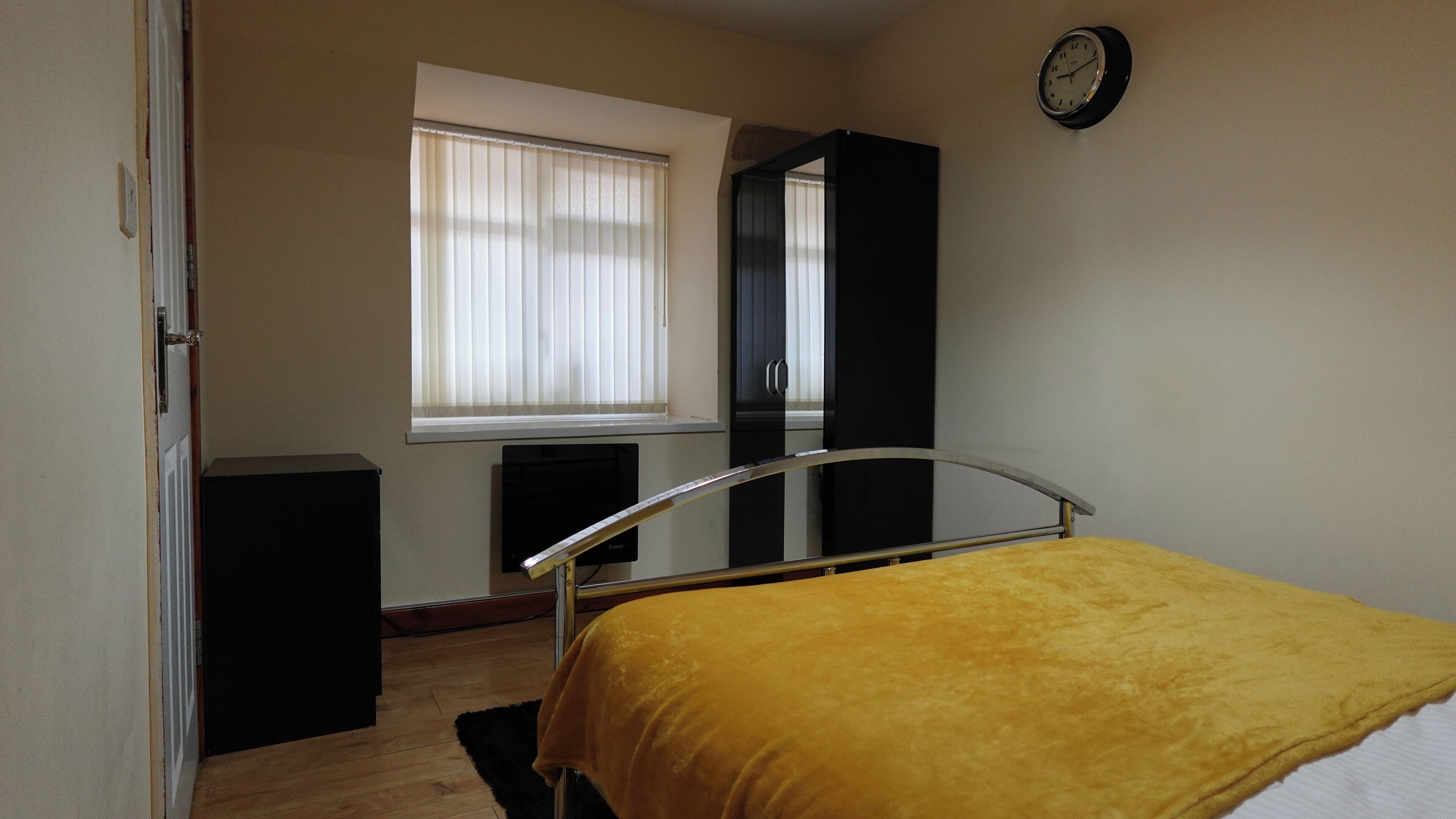 A bedroom with a bed, a window with blinds, a black wardrobe, and a wall clock.