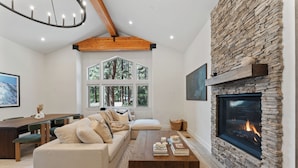 A modern living room with a stone fireplace, a large window with a view of trees, a wooden ceiling, and a cozy seating area. - One Block to Beach, Restaurants, Bars, Brewery. Modern mountain luxury. (Kings Beach)
