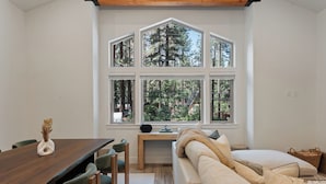 A modern interior with a wooden dining table, green chairs, a beige sofa, and a large window with a view of trees. - One Block to Beach, Restaurants, Bars, Brewery. Modern mountain luxury. (Kings Beach)