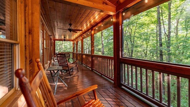 Um deck de madeira com mesa e cadeiras, cercado por guarda-corpo, com vista para uma área arborizada.