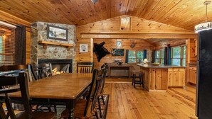 Interior de cabana de tronco com sala de jantar, cozinha e lareira de pedra.