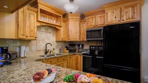 Una cocina con gabinetes de madera, encimeras de granito, un refrigerador negro y diversos alimentos sobre el mostrador.