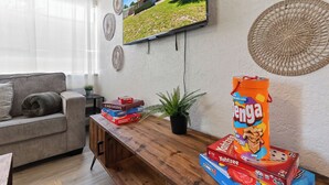 A living room with a sofa, a coffee table with board games, a TV on the wall, and a window with blinds. - Walkable Gem : Retro Arcades : Near Lauderdale-by-the-Sea (Fort Lauderdale)