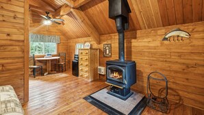Ein gemütliches Blockhaus-Interieur mit Kaminofen, einem Essbereich mit Tisch und Stühlen sowie einem Deckenventilator.