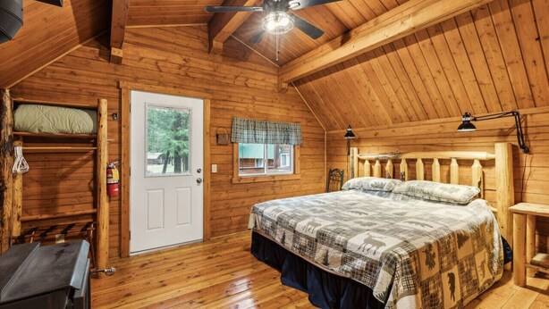 A cozy cabin room with a wooden ceiling, a bed with a plaid bedspread, a window with a plaid valance, and a door with a white frame. - Log Cabin Retreat in the Montana Wilderness (Hamilton)