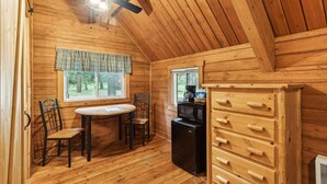 Interior acogedor de una cabaña con techo de madera, ventilador de techo, mesa de comedor con sillas, zona de cocina con microondas y un amplio aparador de madera.
