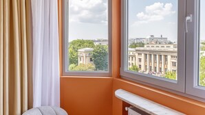 A corner room with an orange wall, a large window with a view of a building, a radiator, and a grey chair with a pillow. - Playful & Bright 1BR near The Faculty of Law (Bucharest)