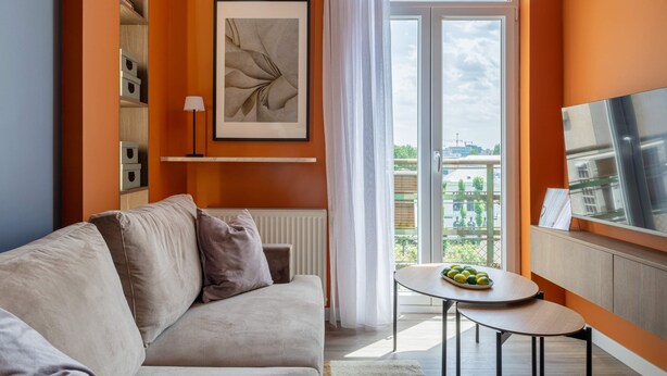 A modern living room with a sofa, a small table with fruit, a TV, and a large window with a view of buildings. - Playful & Bright 1BR near The Faculty of Law (Bucharest)