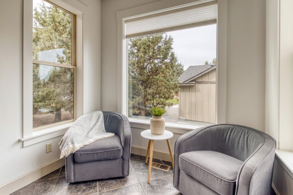 A corner room with two grey armchairs, a small round table with a plant, and large windows with a view of trees and a building.