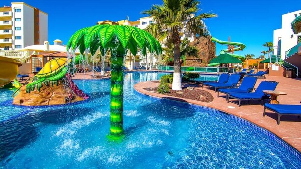 Ein Poolbereich mit einer Palme, einem Wasserspiel, Liegestühlen und einer Wasserrutsche.