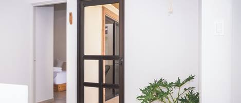 A hallway with a black-framed door leading to a bedroom with a bed and a chair. There is a potted plant on the floor and a hanging plant on the wall.