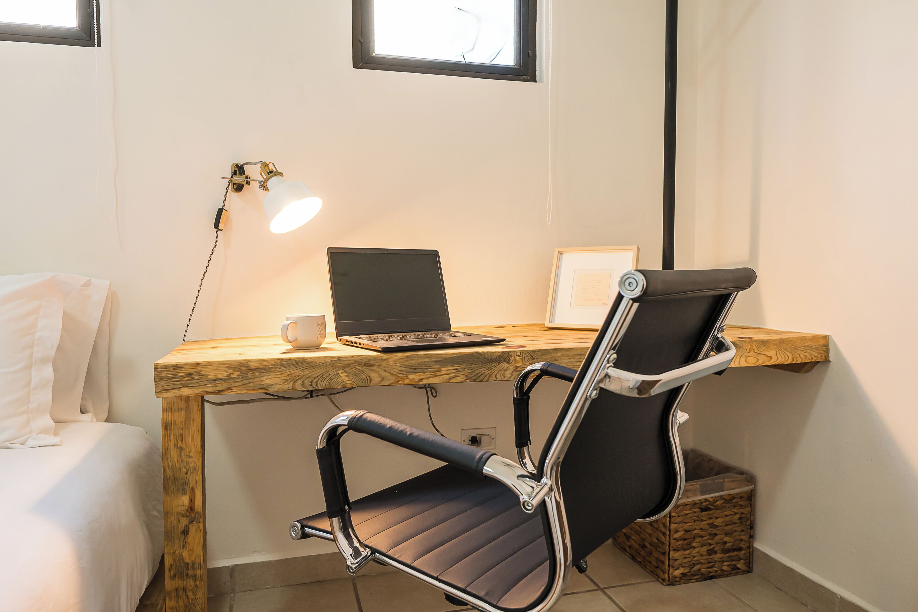 A small, modern room with a wooden desk, a laptop, a chair, and a bedside table with a lamp.