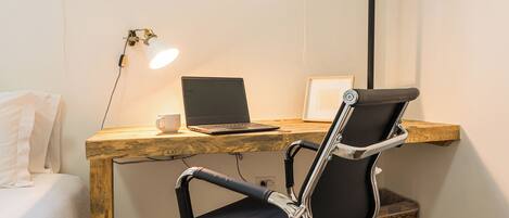 A small, modern room with a wooden desk, a laptop, a chair, and a bedside table with a lamp.