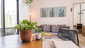 A modern living room with a wooden table, a potted plant, a coffee cup, and a sofa with cushions. There are two wall-mounted paintings and a hanging plant. - Grand Villa w Private Patio & Parking  (San José del Cabo)