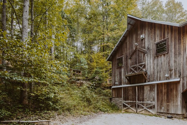 Une grange rustique en bois, avec un sol en terre battue devant, entourée d’arbres denses.