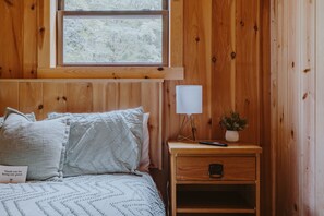 Una habitación de madera con una cama, una mesita de noche, una lámpara y un ventanal.