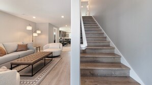 Una sala de estar moderna con un sofá, un reposapiés y una lámpara de pie. Una escalera con peldaños de madera conduce a un piso superior.