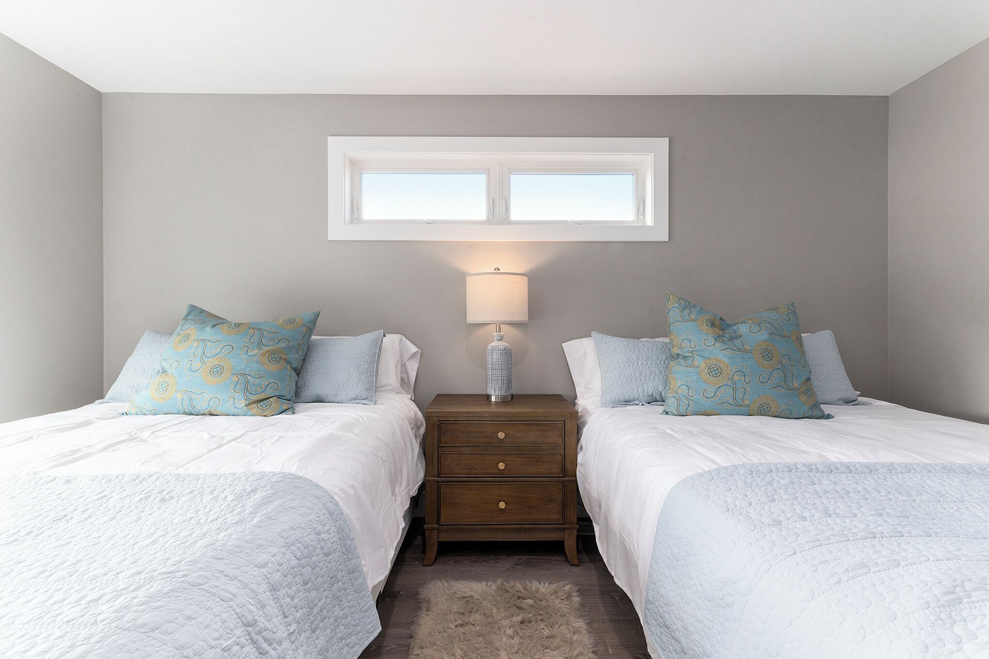 A bedroom with two beds, a nightstand with a lamp, a window above the beds, and a fluffy rug on the floor.