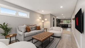 A modern living room with a sofa, armchair, ottoman, and a TV mounted on the wall.