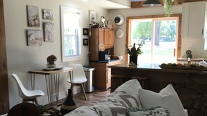 A cozy living room with a sofa, a dining table, and a kitchen area with wooden cabinets and a hanging plant. - Bay Dream Believer Cottage on Beautiful East River Waterfront (Mathews)