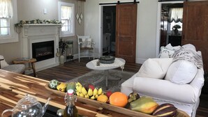 A cozy living room with a fireplace, a white sofa, a wooden coffee table with fruit, and a ceiling fan. - Bay Dream Believer Cottage on Beautiful East River Waterfront (Mathews)