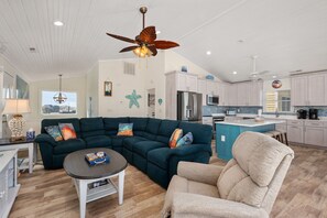 Amplia sala de estar con un sofá modular, una mesa de centro y un sillón color beige. Se aprecia una cocina con armarios blancos y una isla azul al fondo.