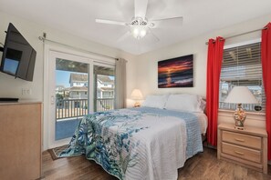 Une chambre avec un lit, un ventilateur de plafond, un téléviseur fixé au mur, une fenêtre avec des rideaux rouges et un balcon visible à travers la fenêtre.