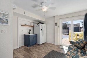 Une cuisine compacte équipée d’un réfrigérateur, d’un évier et de meubles en bois. On y trouve également un ventilateur de plafond et une porte-fenêtre coulissante qui donne sur un espace extérieur avec une table et des chaises jaunes.