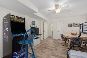 Une pièce équipée d’une console de jeux, d’une table à manger avec des chaises, d’un ventilateur de plafond et d’une porte donnant sur une autre pièce.