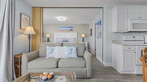 Un salón con un sofá, una mesa de centro de vidrio con pasteles, una cocina con armarios blancos y un microondas, y un cuadro con motivos de playa en la pared.