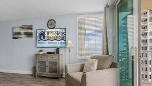 Una sala de estar con un sofá color beige, un televisor mostrando 'Relens Vacations', un reloj y una ventana con persianas.