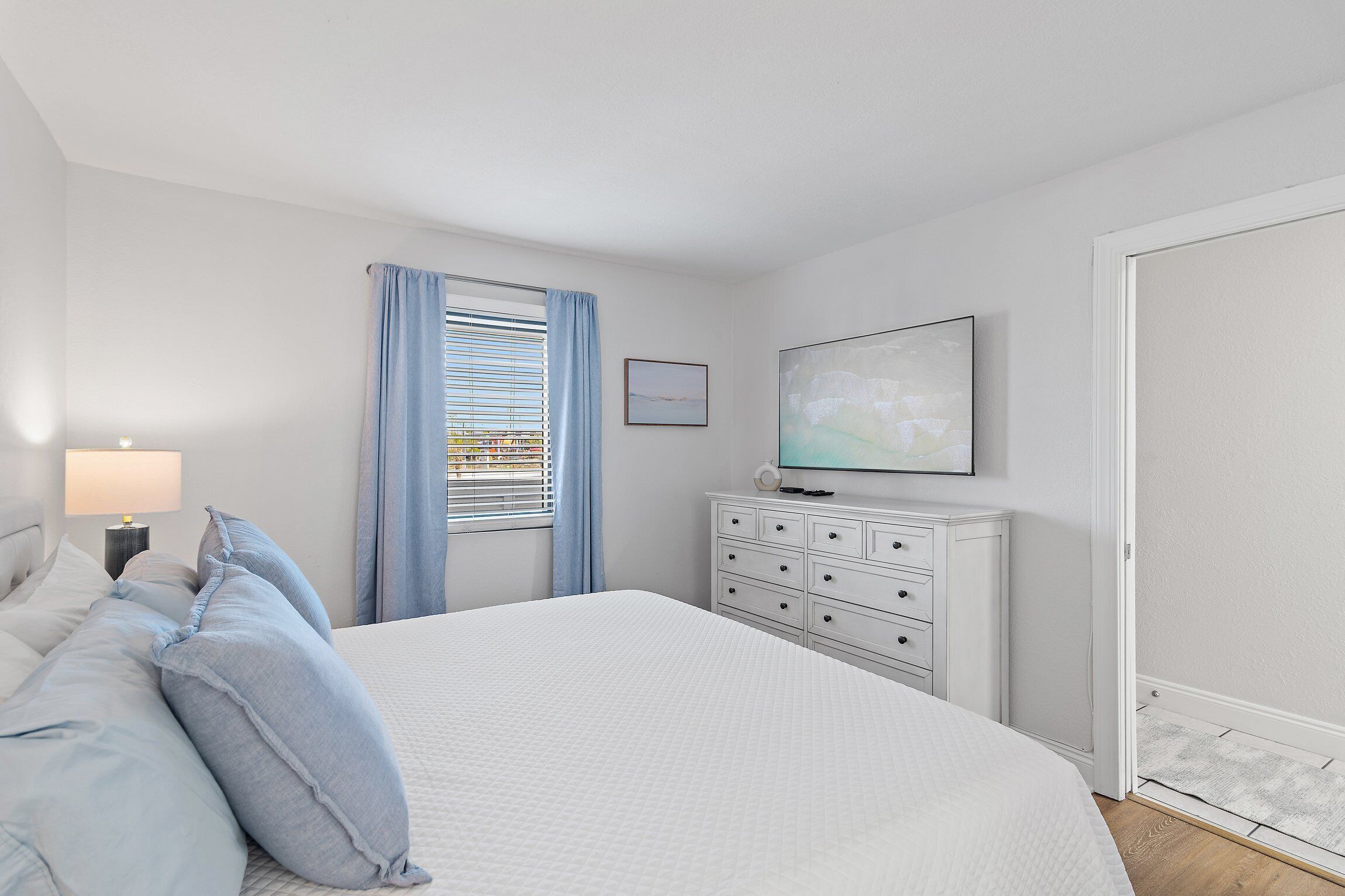 A bedroom with a large bed, a dresser, a nightstand, a lamp, a window with curtains, and a painting on the wall.