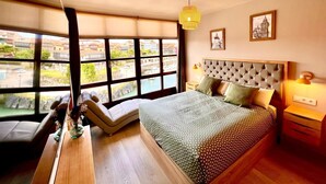 A bedroom with a large window, a bed with a tufted headboard, bedside lamps, and a view of buildings outside.